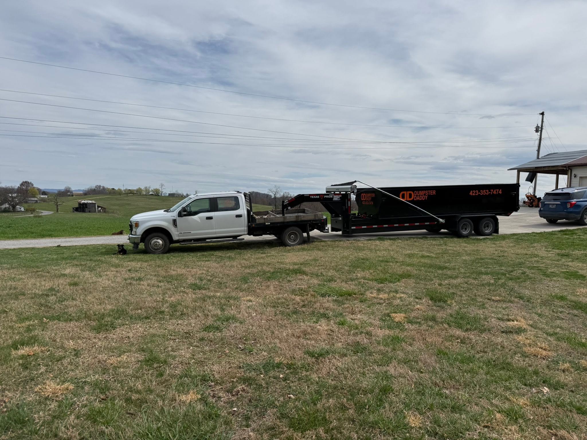 White pickup truck towing a Dumpster Daddy dumpster trailer on a rural property